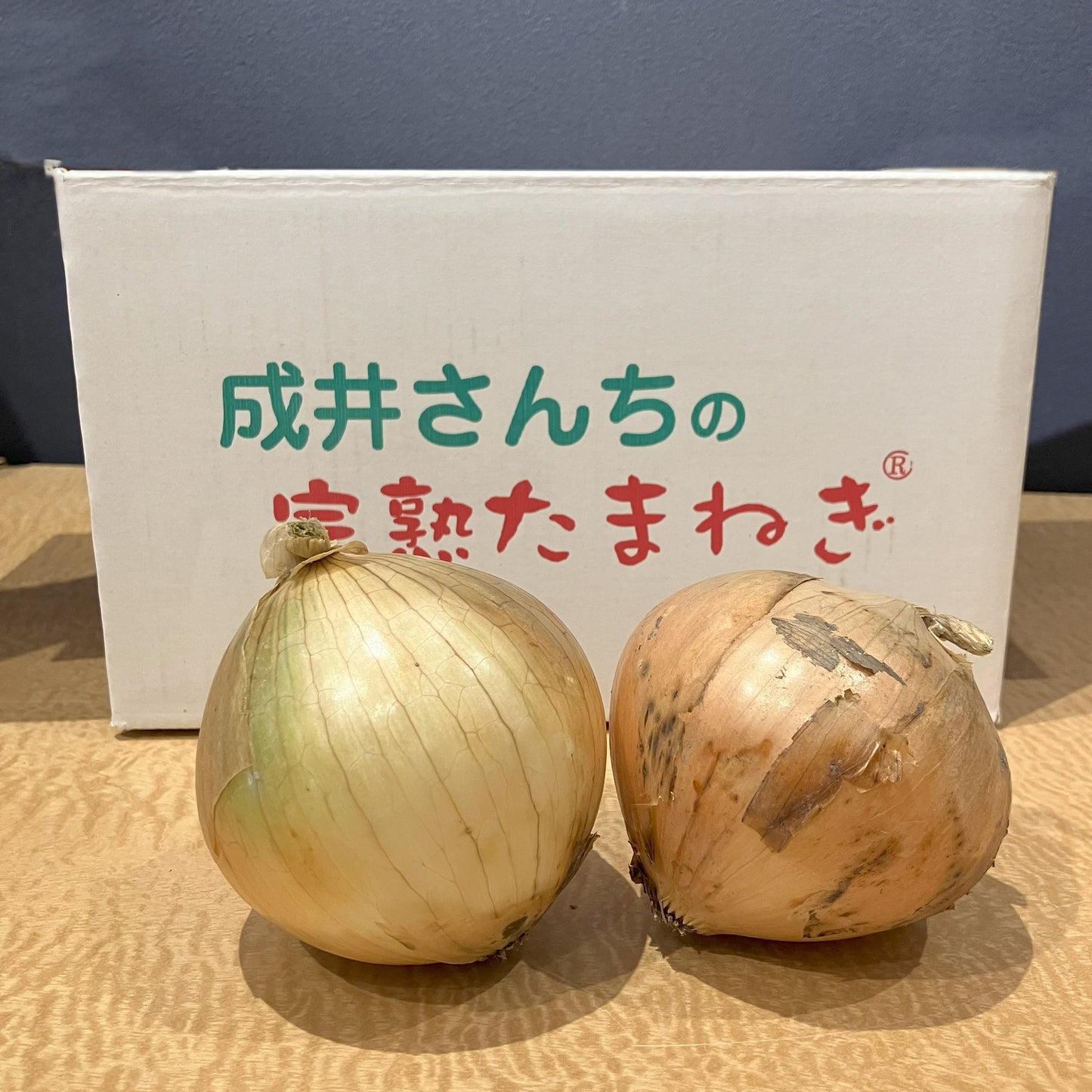 成井さんちの完熟たまねぎのロゴが入った段ボールの前にたまねぎを2個並べています