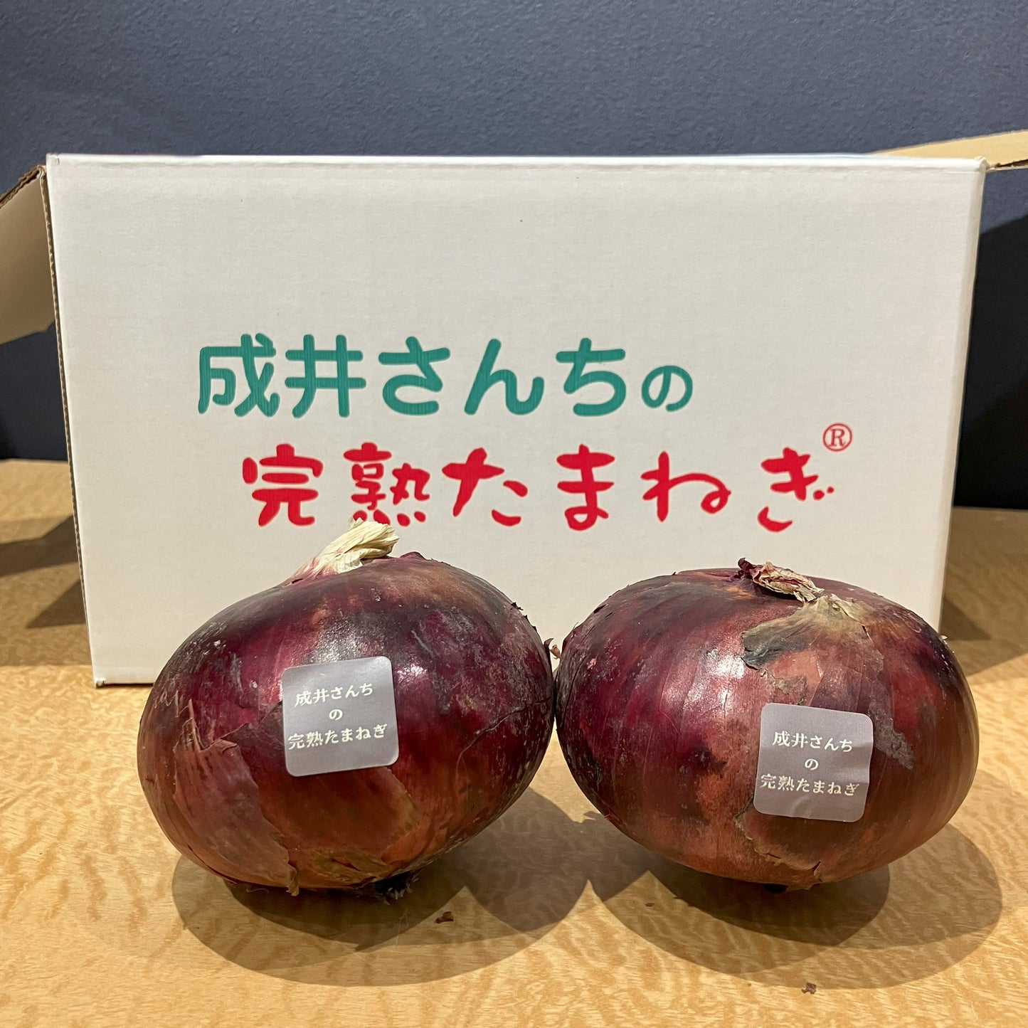 成井さんちの完熟たまねぎ（甘丸ルビー）1kgは、じっくり育てられた赤玉ねぎで、アントシアニンがたっぷり詰まっています。ナスや紫イモ、紫キャベツにも負けないこの紫色のポリフェノールは、体の中の活性酸素をやっつけて、強力な抗酸化パワーを発揮！生活習慣病の予防や血のめぐりアップにも役立ち、元気な毎日を応援します。鮮やかな色と栄養満点の自慢の一品です。成井さんち専用の箱の前に玉ねぎ２つ置いた写真
