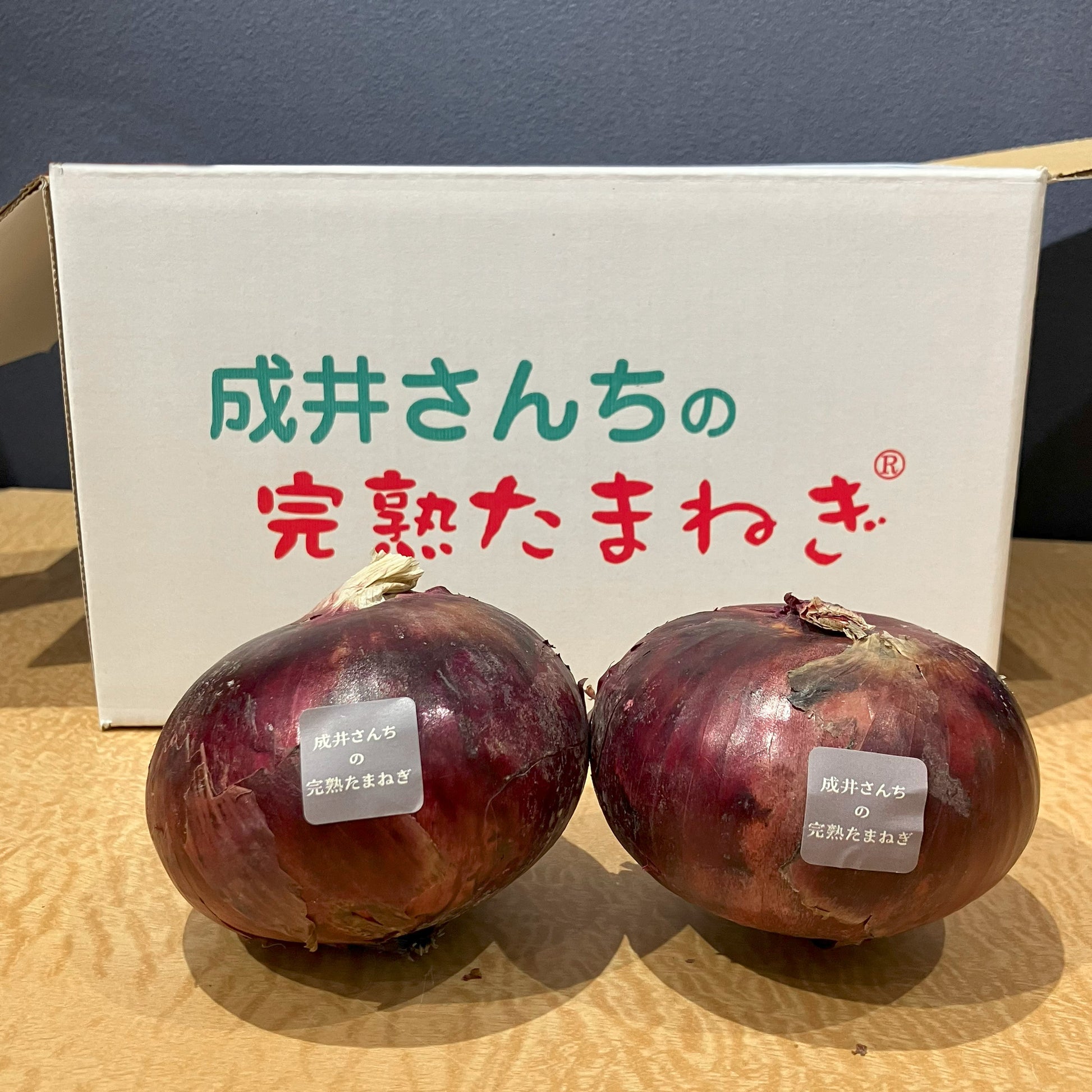 成井さんちの完熟たまねぎ（甘丸ルビー）1kgは、じっくり育てられた赤玉ねぎで、アントシアニンがたっぷり詰まっています。ナスや紫イモ、紫キャベツにも負けないこの紫色のポリフェノールは、体の中の活性酸素をやっつけて、強力な抗酸化パワーを発揮！生活習慣病の予防や血のめぐりアップにも役立ち、元気な毎日を応援します。鮮やかな色と栄養満点の自慢の一品です。成井さんち専用の箱の前に玉ねぎ２つ置いた写真