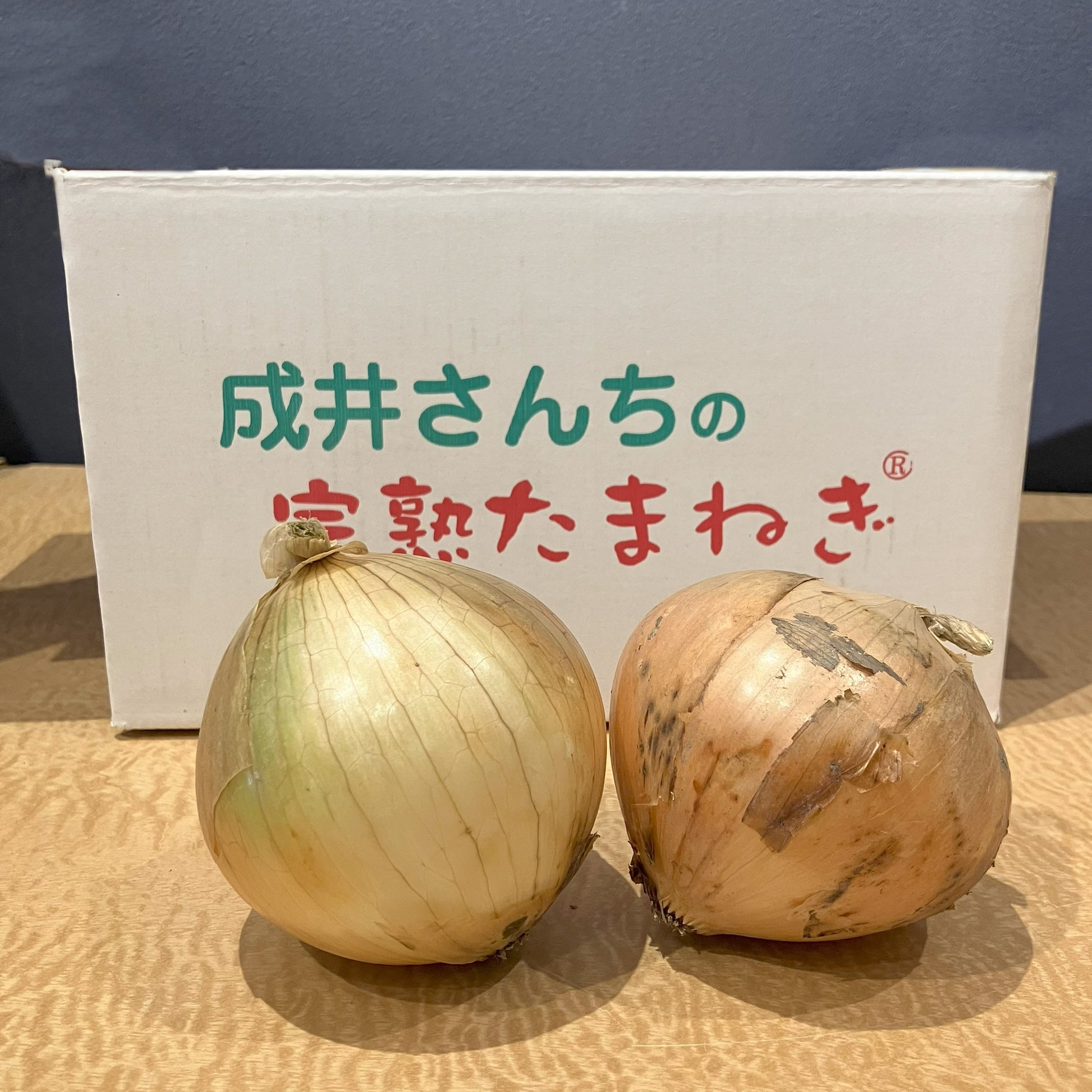 成井さんちの完熟たまねぎのロゴが入った段ボールの前にたまねぎを２個並べています